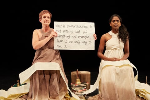 Sue MacLaine holds a hand-lettered placard reading 'What is incomprehensible is that nothing, and yet everything has changed. That is the only way to put it,' beside a calm, seated Nadia Nadarajah.