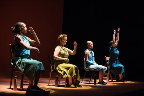 The four performers of Vessel, seated on a red-lit stage, raise their hands and arms together in a synchronised gesture.