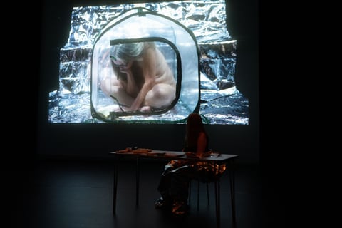 Sue MacLaine sits with her back to us at a foil-strewn table on a dark stage, facing a large projection of a naked, grey-haired figure curled inside a glowing translucent pod amid crumpled silver foil.