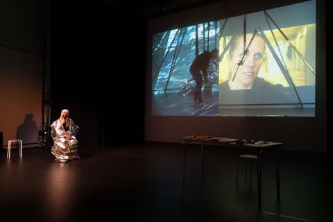 Wrapped in a silver foil blanket, Sue MacLaine kneels beside a small table on a dark stage as a large projection shows a figure climbing an icy slope alongside a close-up of her face.