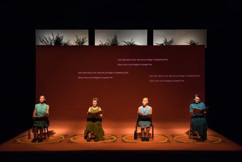 Four performers sit on chairs, each in a pool of light on a deep-red stage, beneath projected text reading 'Let's talk about who has the privilege of speaking first.'