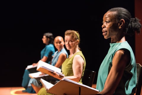 Four women in sleeveless green and teal dresses sit in a row on a darkened stage, each holding an open spiral-bound script; the nearest, in teal, speaks in profile.