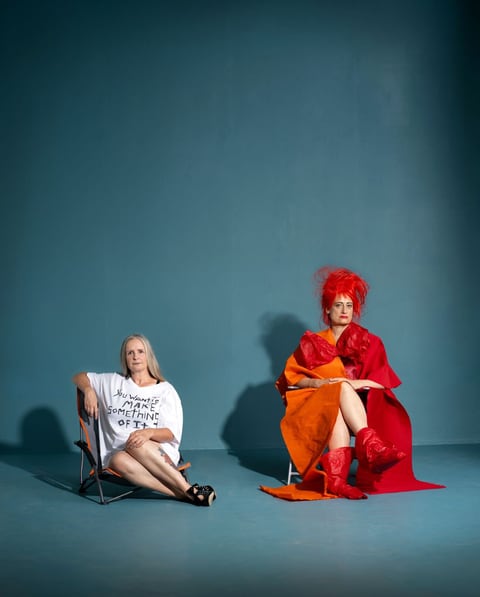 Against a teal backdrop, Sue MacLaine sits in an oversized hand-lettered t-shirt reading 'You want to make something of it', beside Hannah Ringham in a flamboyant red-and-orange costume with spiked red hair.